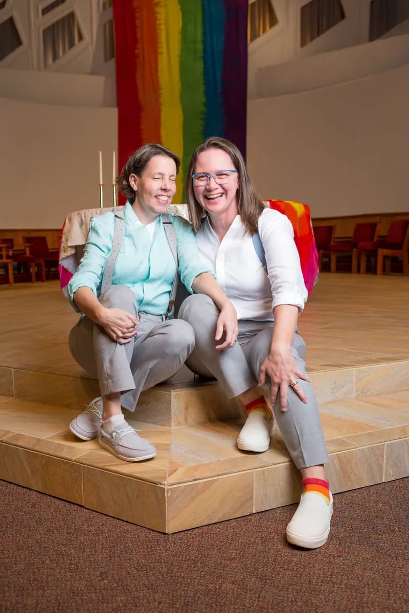 Two brides laughing together on the altar steps in front of a rainbow flag